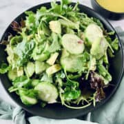 French green salad in a black bowl with a mint linen napkin at the bottom and dressing in a small black bowl in the top right corner.