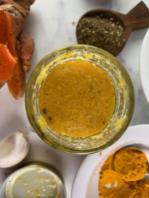 OVERHEAD VIEW OF SHAKEN TURMERIC DRESSING IN A GLASS JAR WITH TURMERIC, GARLIC, SALT AND PEPPER ON THE SIDE