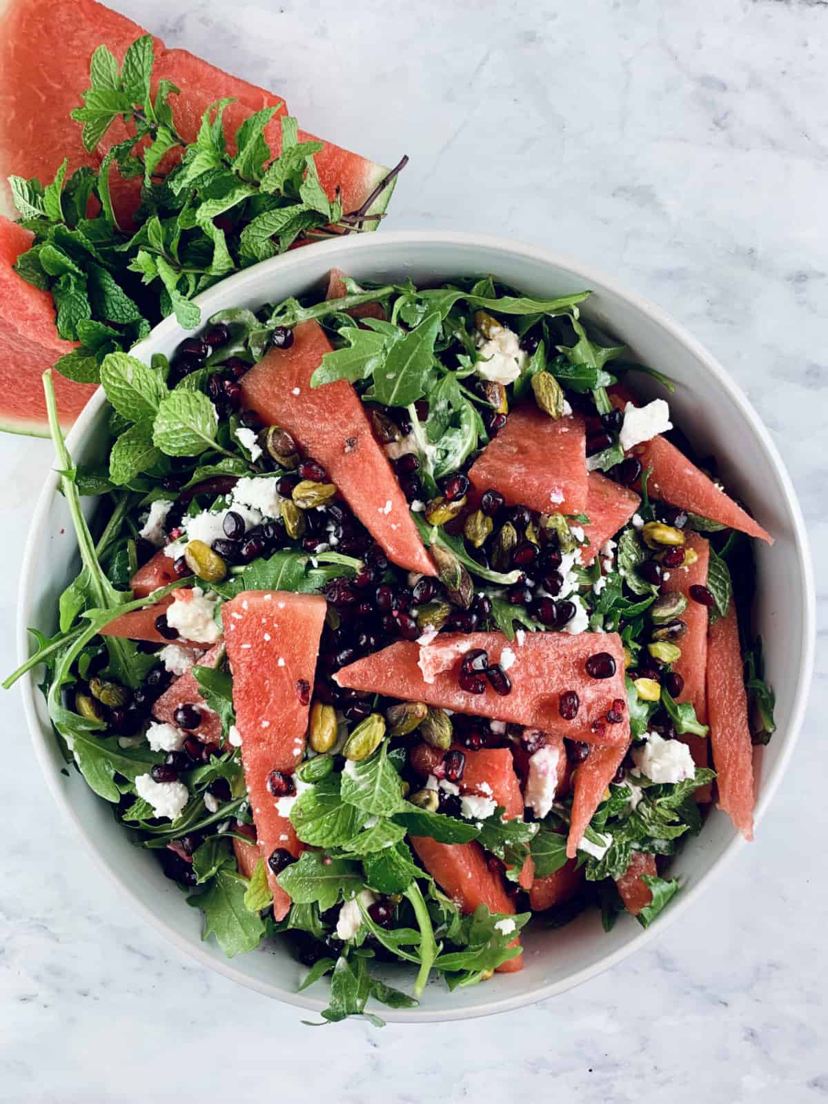 CLOSE UP OF PERSIAN WATERMELON & FETA SALAD IN A WHITE BOWL WITH WATERMELON SLICES & MINT ON THE SIDE