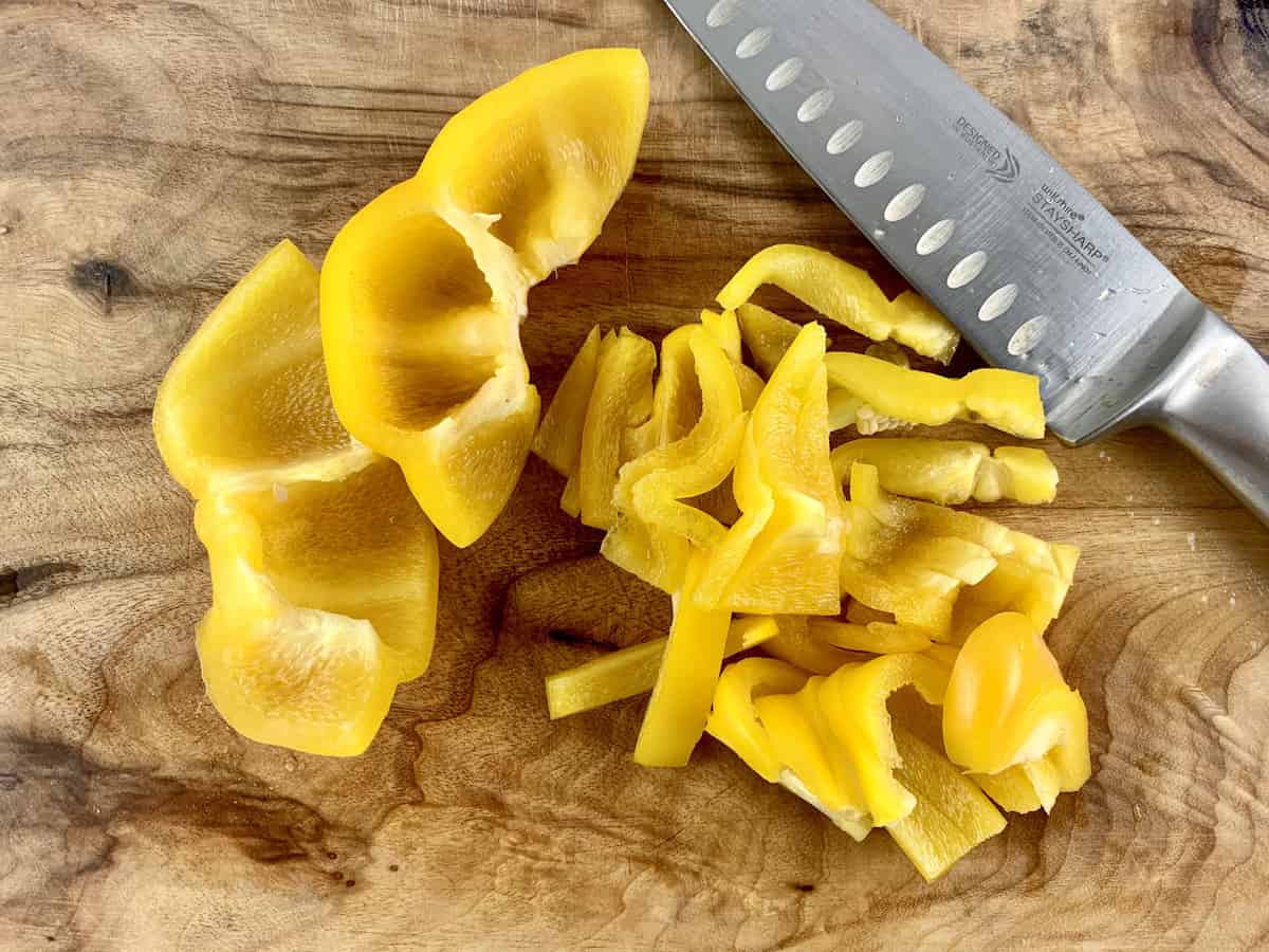 Yellow peppers that have been cut into strips and in half on a wooden board with a knife on the side.