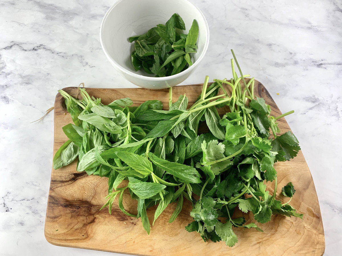 Picking basil, Vietnamese mint & coriander leaves from stems on a wooden board with a white bowl in the background from stems.