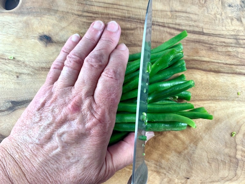 HANDS LINING UP TO CUT GREEN BEANS IN HALF WITH A KNIFE ON A WOODEN BOARD