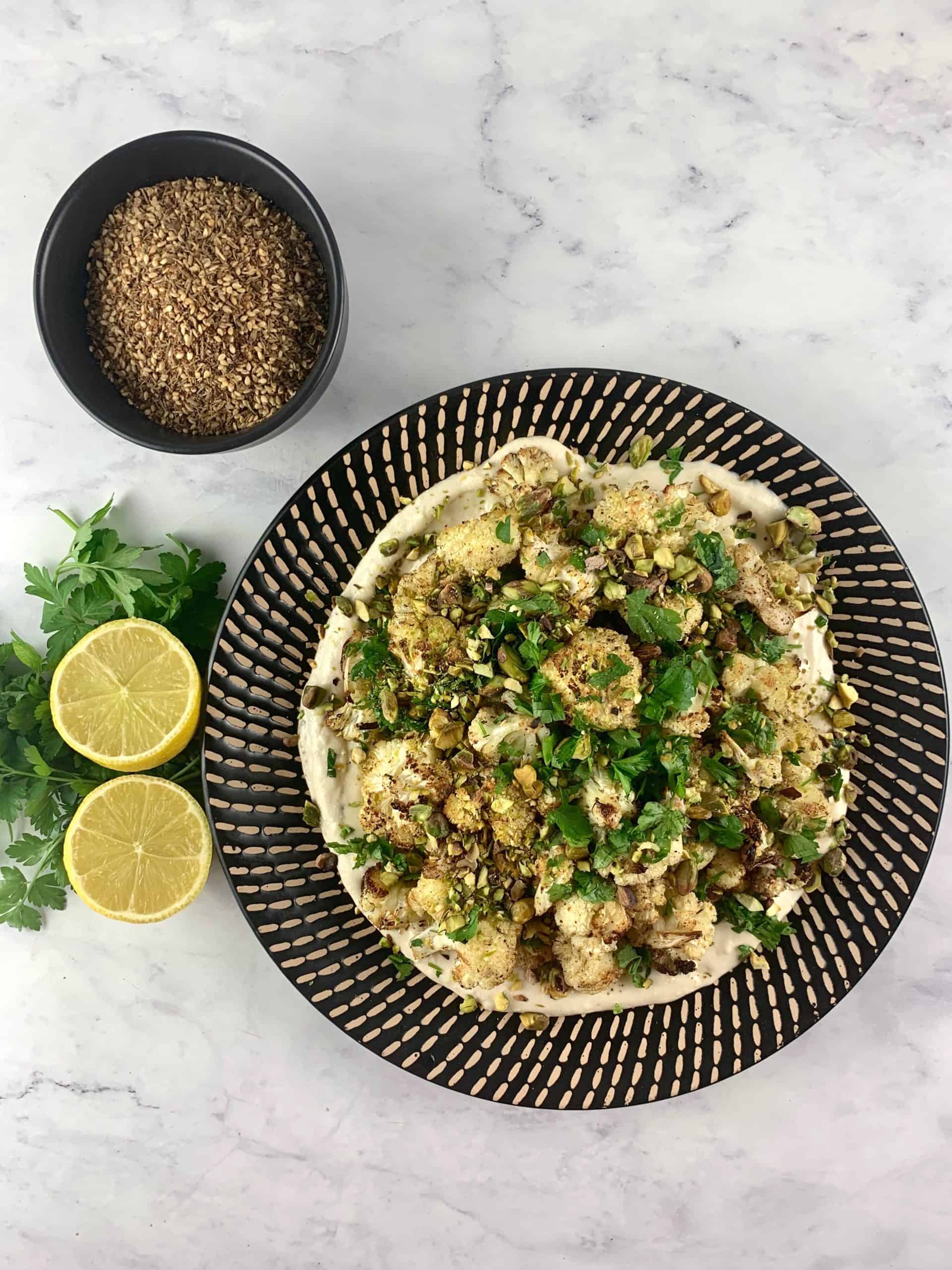 CAULIFLOWER TAHINI SALAD ON A BLACK PATTERNED PLATE WITH ZA'ATAR AND LEMONS & PARSLEY ON THE SIDE