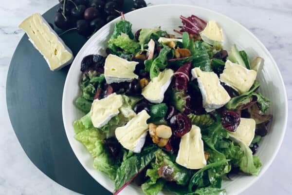 Brie salad with cherries in a white bowl on a dark grey board with cherries and cut brie in top left corner.