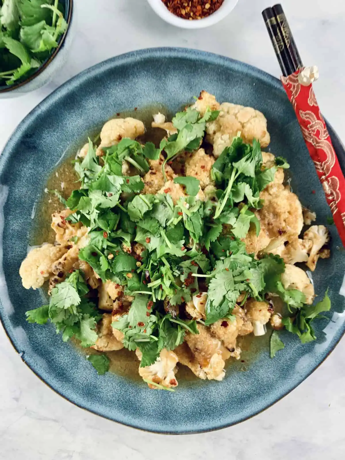 Miso cauliflower in blue bowl with chopsticks, chilli flakes and coriander on the side.