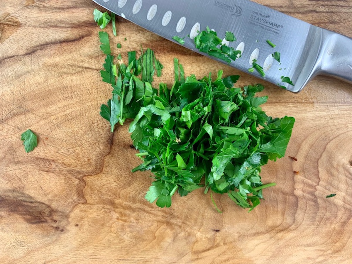 Roughly chopped parsley leaves on a wooden board with a stainless steel knife on the side.