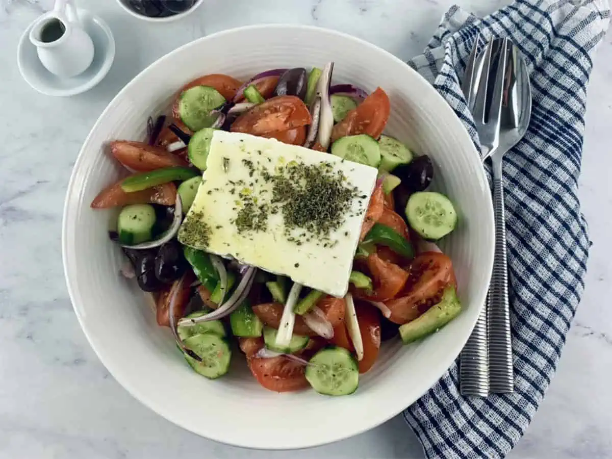 HORIATIKI SALATA IN A WHITE BOWL WITH OIL AND OLIVES ON THE SIDE