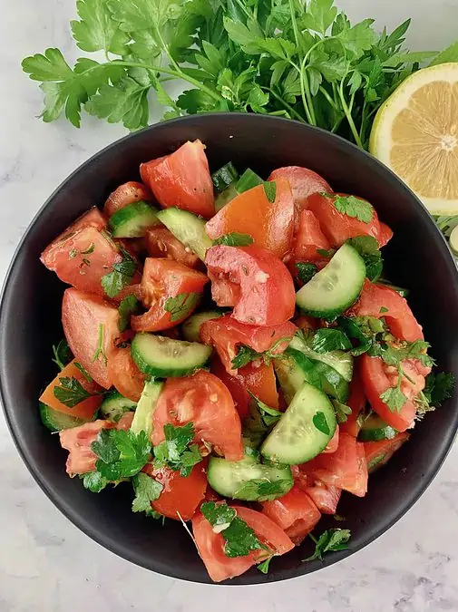 Mediterranean tomato cucumber salad in a black bowl with parsley, lemon and garlic on the side.