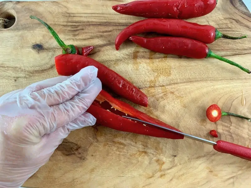 SLICING CHILLIS IN HALF LENGTHWISE ON A WOODEN BOARD WITH A RED KNIFE