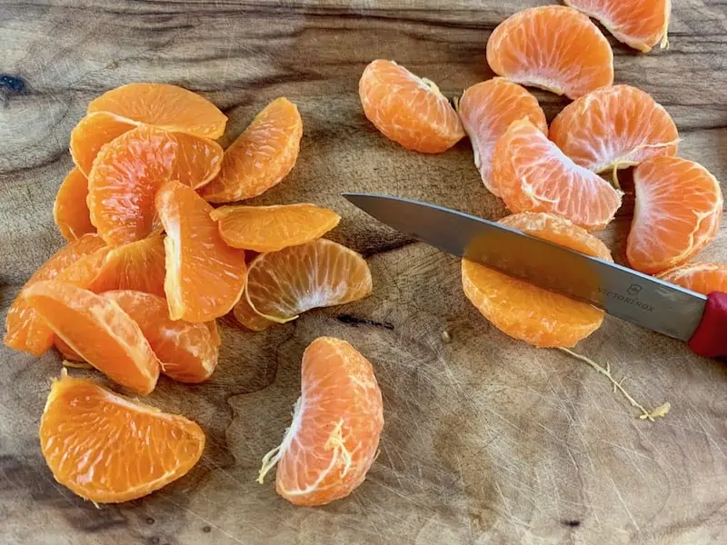 SLICING PEELED MANDARINS LENGTHWISE ON WOODEN BOARD WITH RED KNIFE