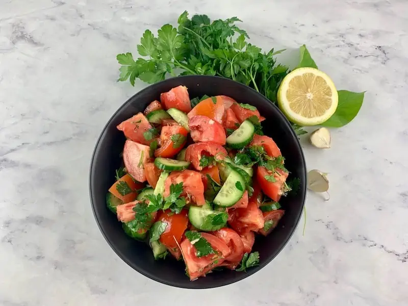 Mediterranean tomato cucumber salad in a black bowl with herbs, lemon and garlic on the side. 