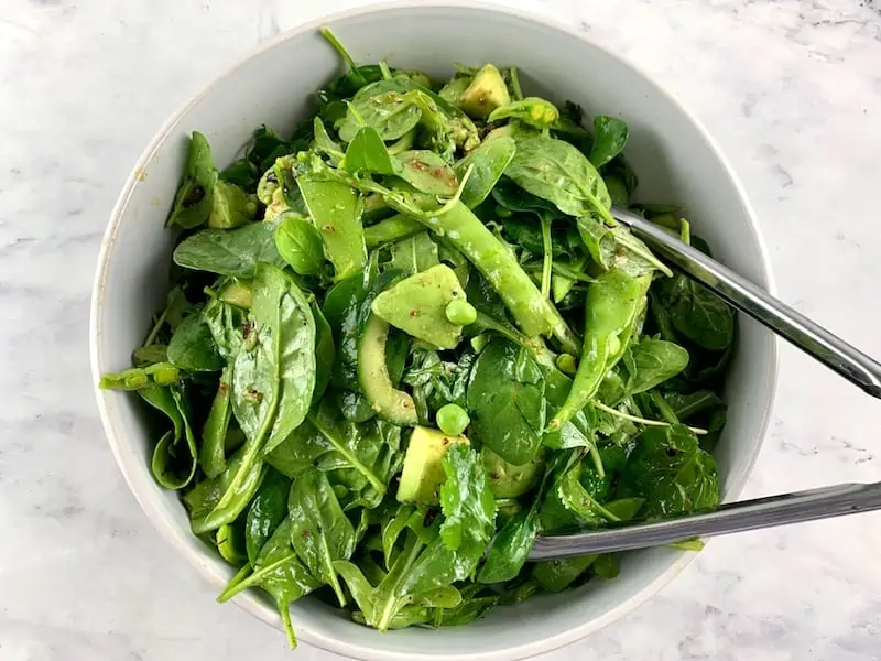 Tossing spinach arugula salad in a white bowl with tongs.
