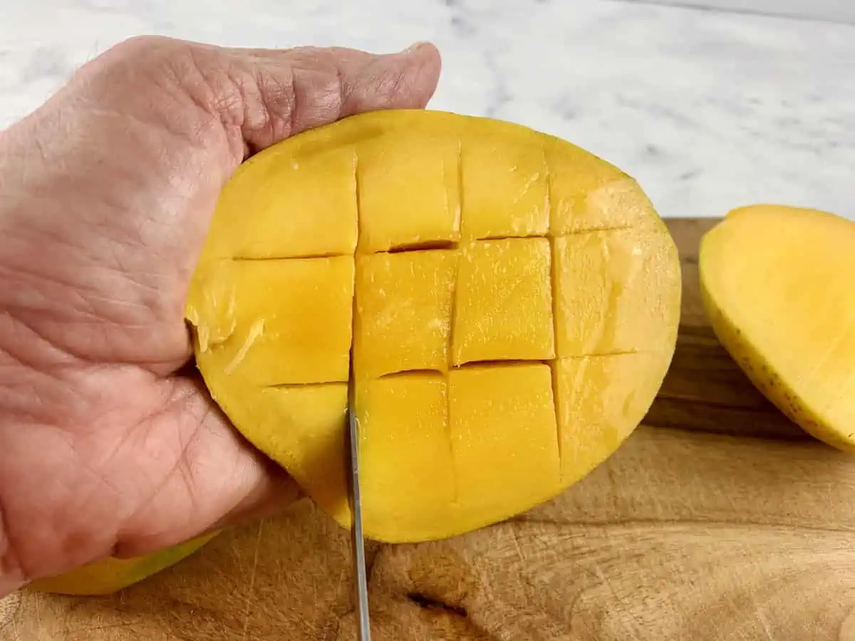 Hands are cutting mango flesh into a cross-hatch pattern with a knife on a wooden board.