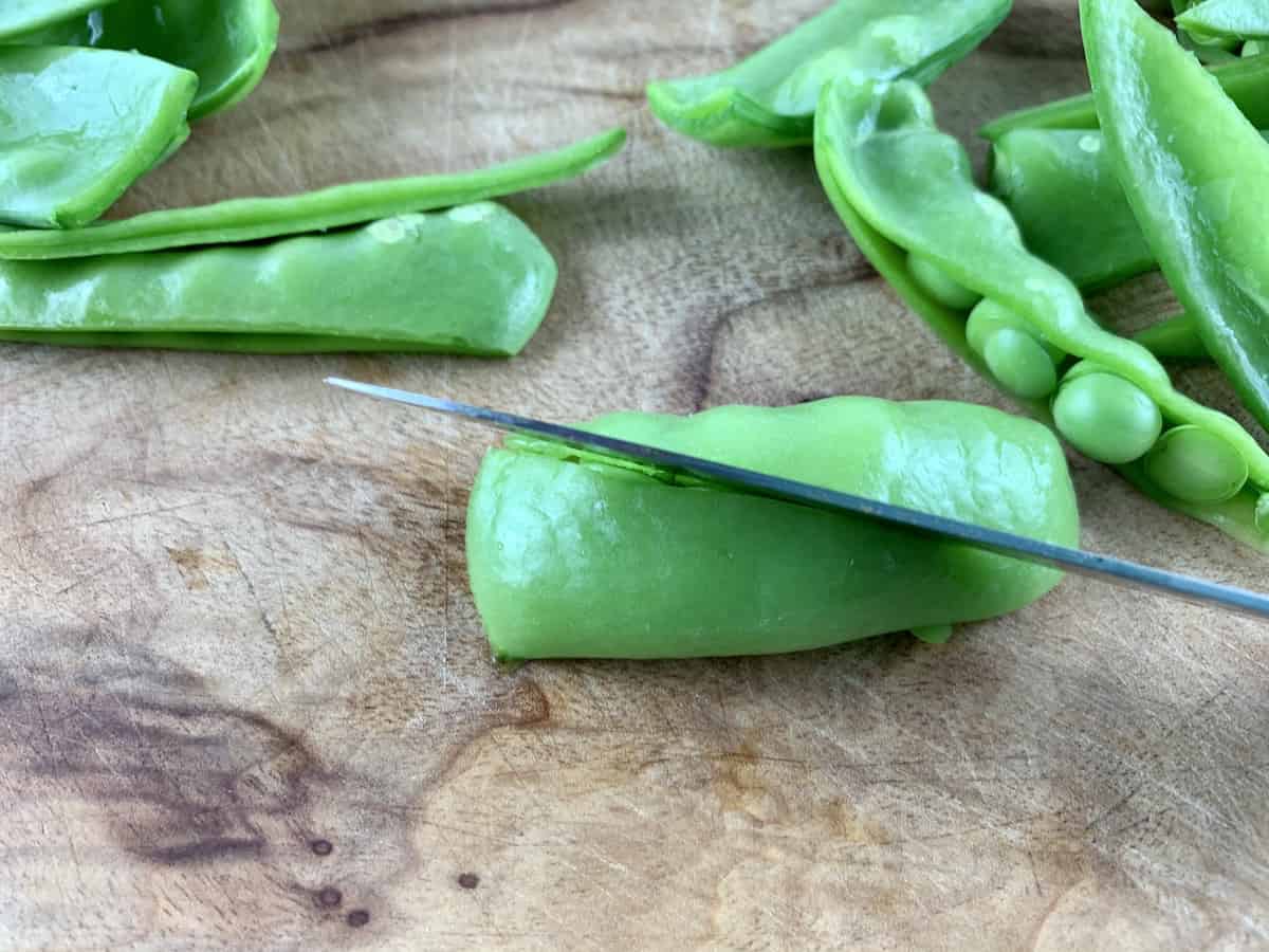 Cutting snow peas diagonally on a wooden board with a knife.