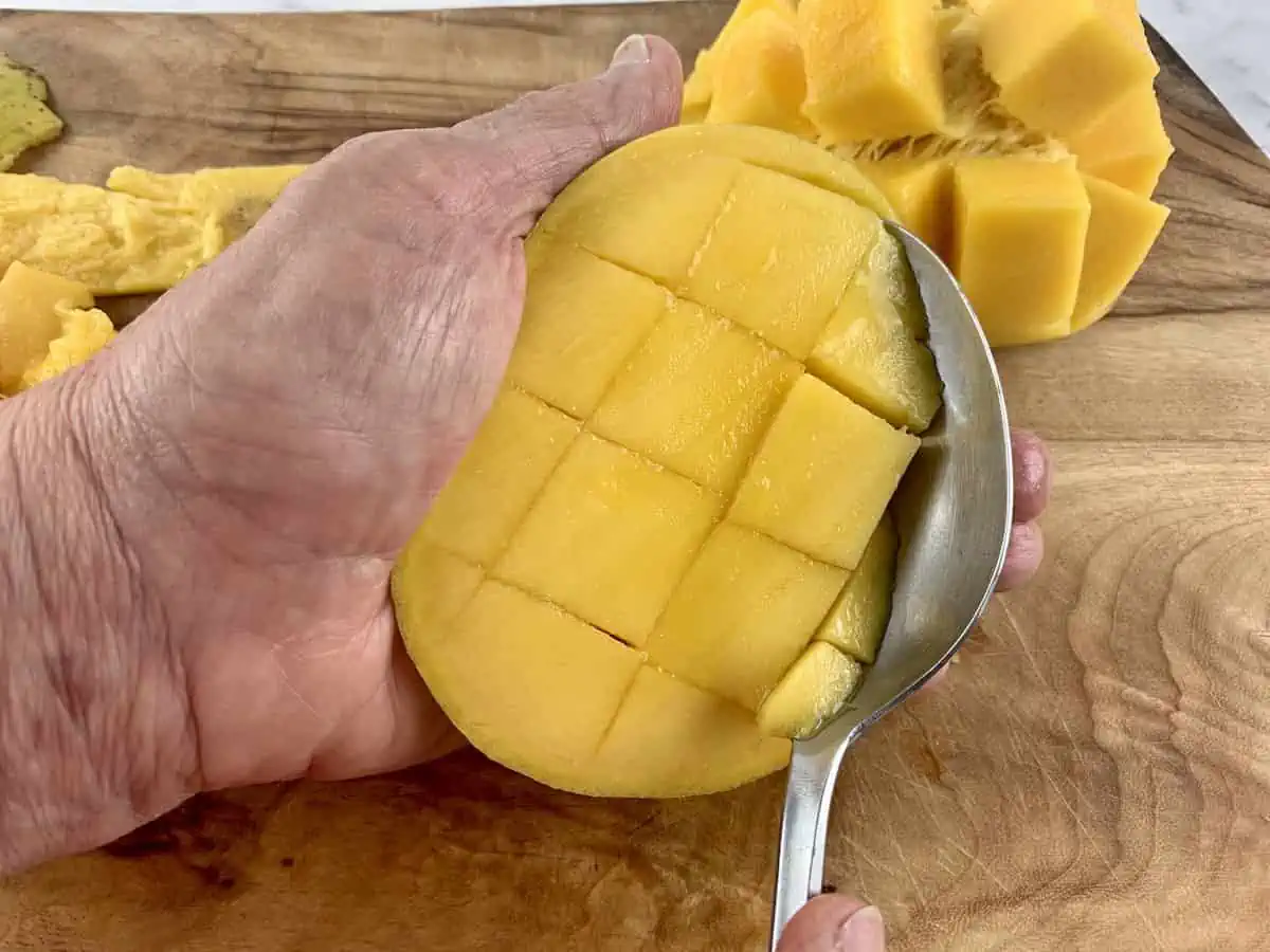 HANDS SCOOPING OUT MANGO FLESH WITH A SPOON ON A WOODEN BOARD