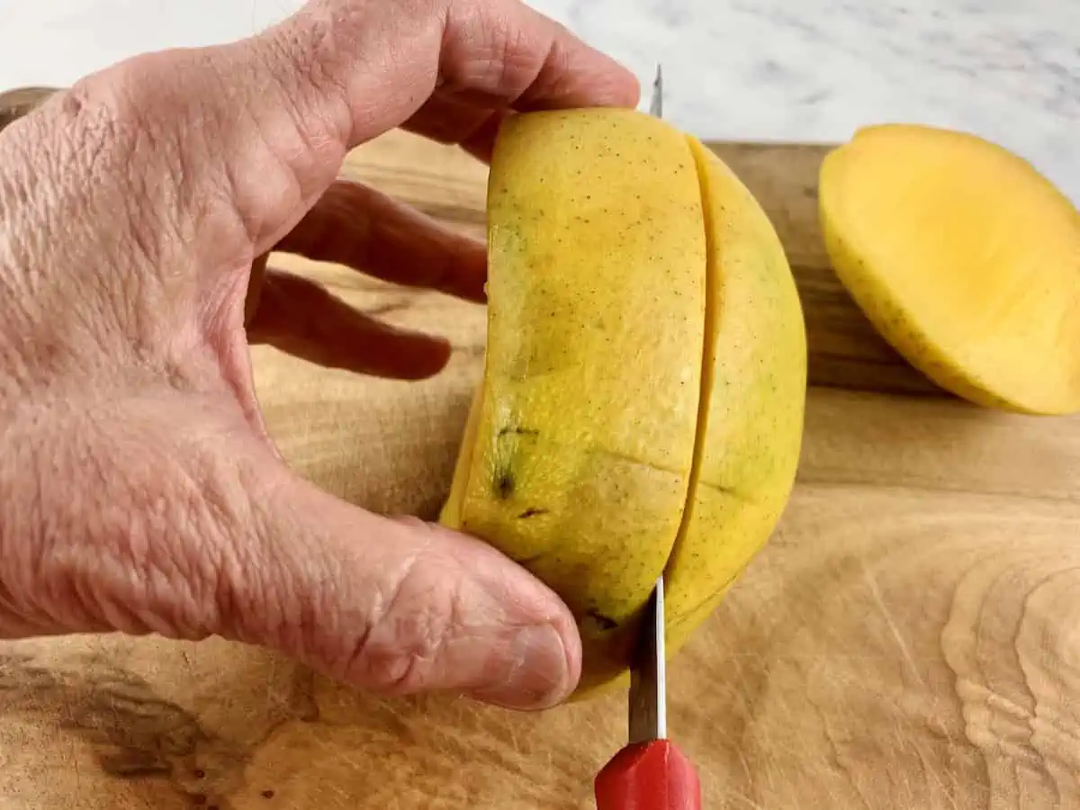 HANDS HOLDING MANGO AND SLICING 2ND CHEEK WITH A KNIFE ON A WOODEN BOARD