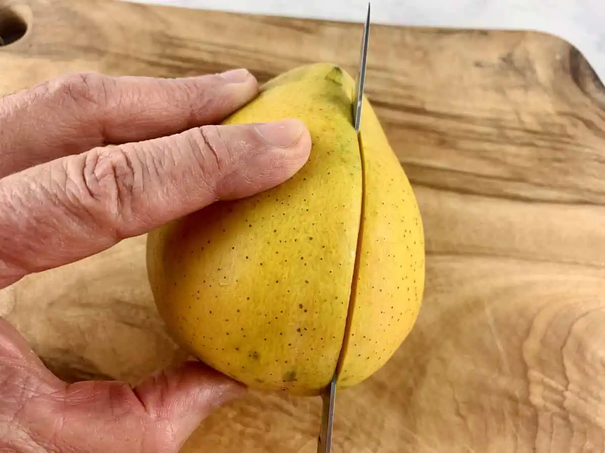 HANDS HOLDING MANGO AND SLICING CHEEK WITH A KNIFE ON A WOODEN BOARD