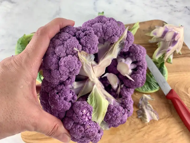 HANDS REMOVING THE CORE FROM PURPLE CAULIFLOWER ON A WOODEN BOARD WITH RED KNIFE ON SIDES
