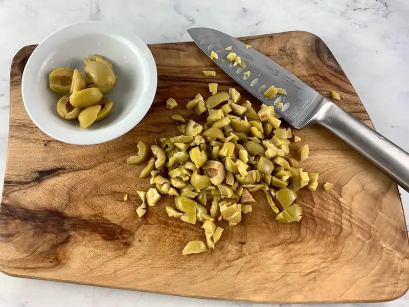 CHOPPED GREEN OLIVES ON A WOODEN BOARD WITH A KNIFE AND HALVED OLIVES IN A WHITE DISH