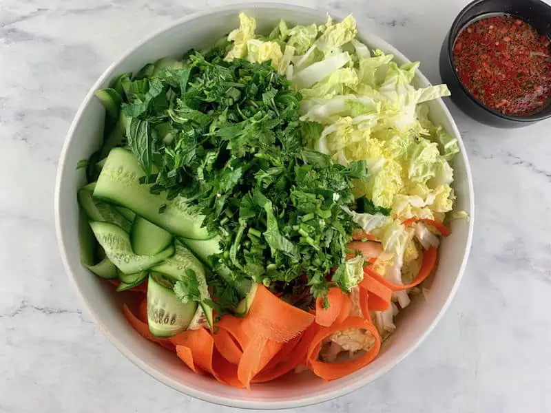 ADDING SALAD INGREDIENTS TO WHITE BOWL WITH DRESSING IN BACKGROUND