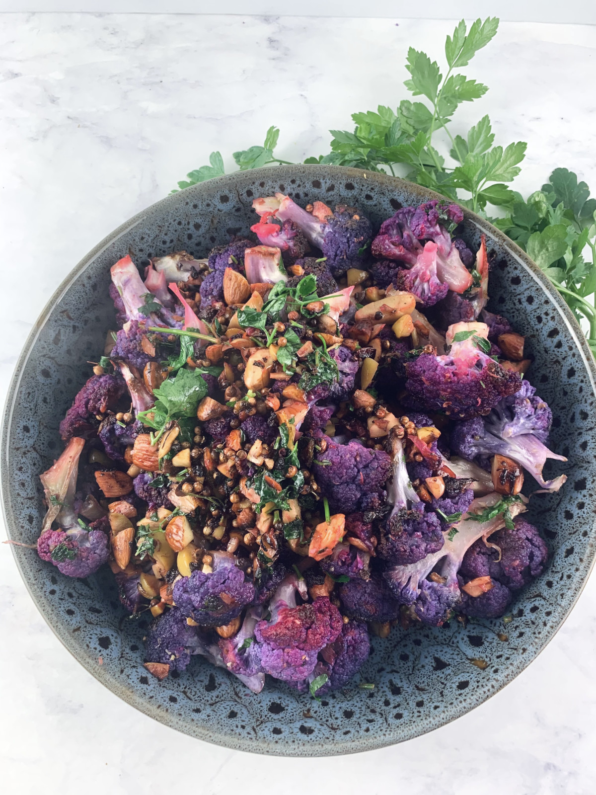 Purple cauliflower salad in shallow dark grey patterned salad bowl with parsley sprigs on the top right corner.