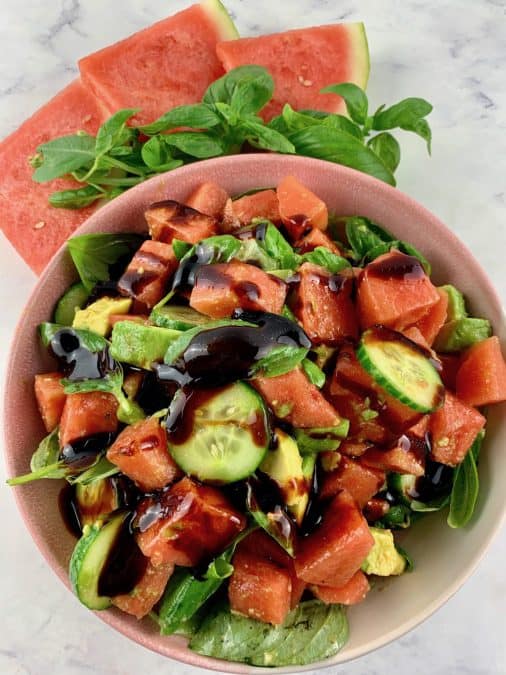 WATERMELON AVOCADO SALAD WITH BALSAMIC GLAZE IN A PINK BOWL WITH WATERMELON SLICES & BASIL ON THE SIDE