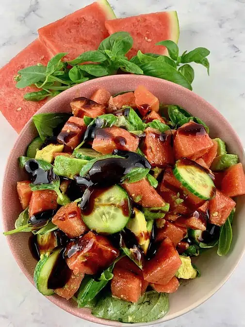 WATERMELON AVOCADO SALAD WITH BALSAMIC GLAZE IN A PINK BOWL WITH WATERMELON SLICES & BASIL ON THE SIDE