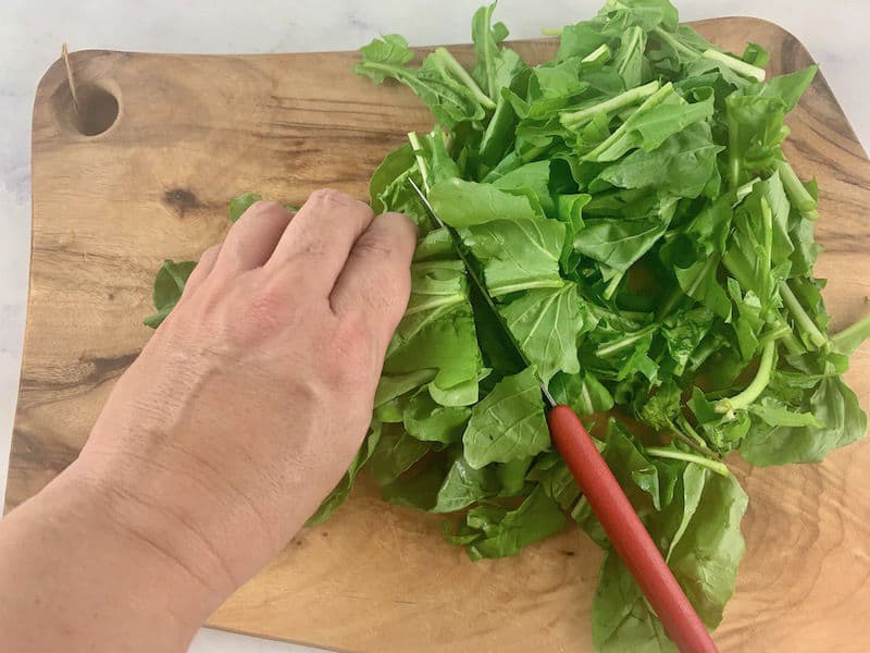 HANDS CHOPPING ARUGULA ON A WOODEN BOARD WITH A RED KNIFE