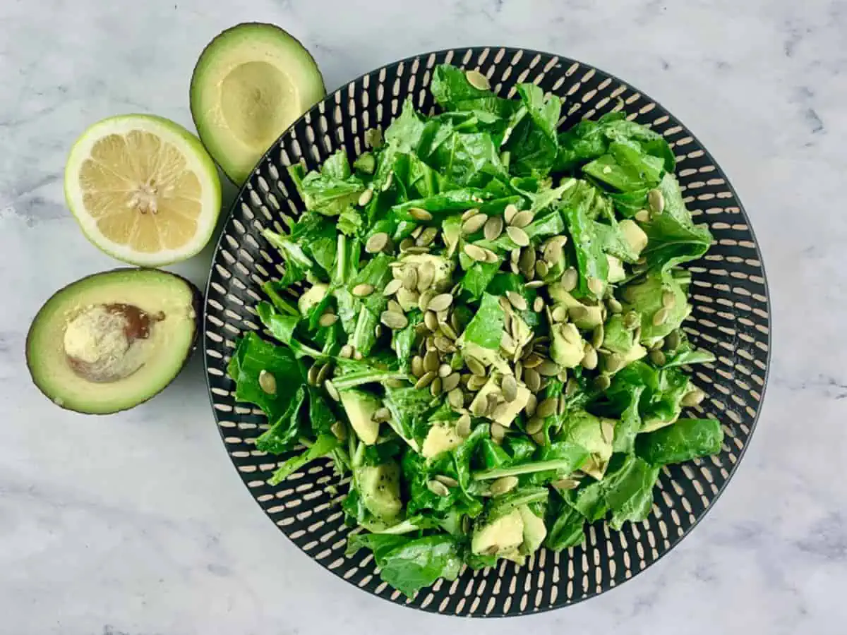 Arugula avocado salad on black patterned plate with avocado halves and lemon on the side.