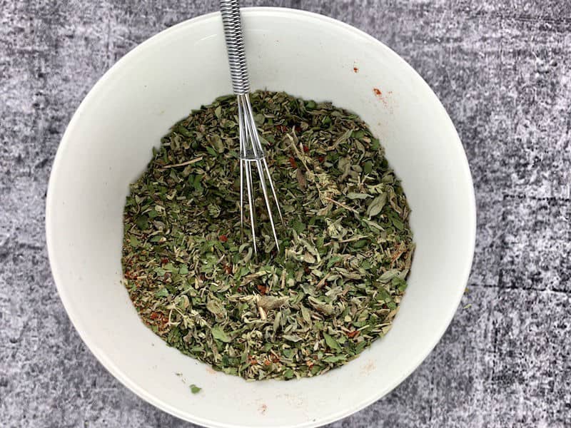 Mixing Italian herb seasoning in a white bowl with a small whisk.