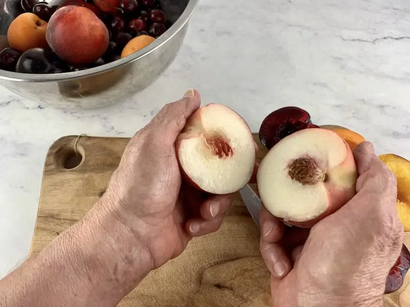 HANDS SEPARATING WHITE PEACH WITH HALVED STONE FRUIT ON A WOODEN BOARD IN BACKGROUND