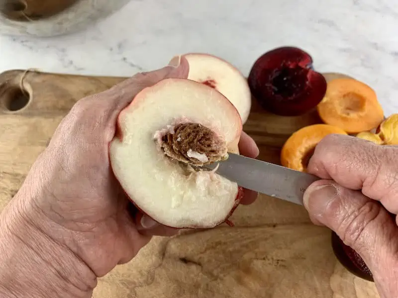 HANDS CUTTING AROUND PIT OF WHITE PEACH WITH HALVED STONE FRUIT ON A WOODEN BOARD IN BACKGROUND