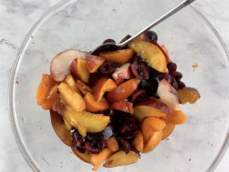 MIXING STONE FRUIT SALAD WITH VANILLA BEAN SYRUP IN A BOWL WITH METAL SPOON