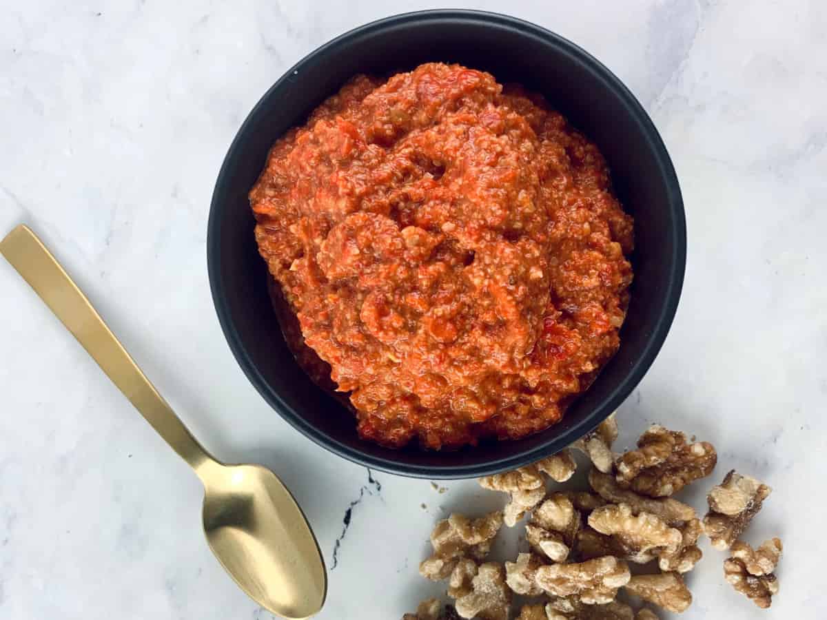 Muhammara dip in black bowl with gold spoon and walnuts on the side.