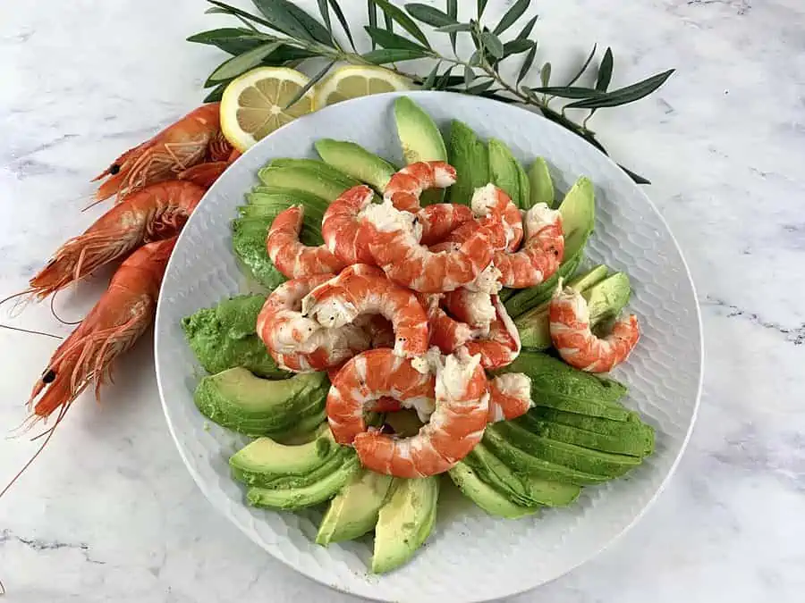 Prawn avocado salad on a white plate with prawns and lemon slices on the side.