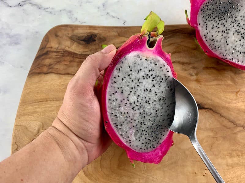 HANDS SCOOPING OUT DRAGON FRUIT FLESH WITH A SPOON ON A WOODEN BOARD