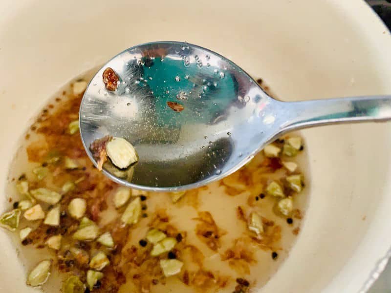 CLOSE UP OF SPOON WITH CARDAMOM SYRUP WITH PAN IN THE BACKGROUND