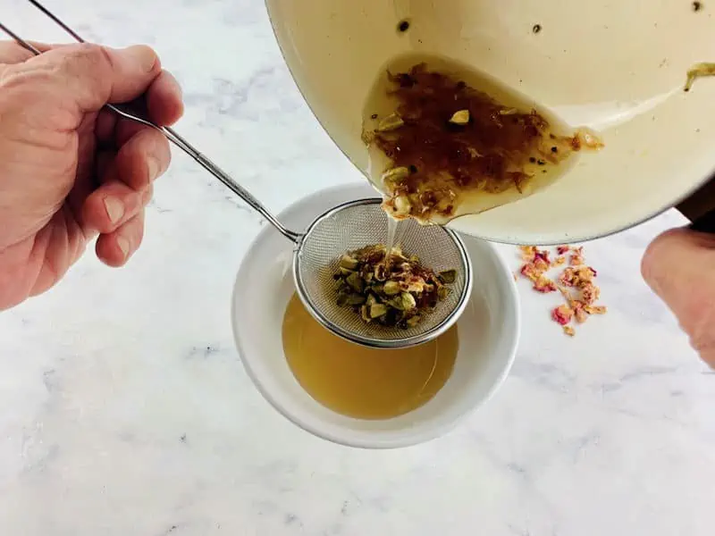 HANDS STRAINING CARDAMOM SYRUP INTO A WHITE BOWL