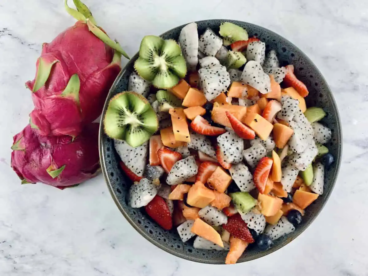 Dragon fruit salad in dark grey bowl with dragon fruit, lime & ginger on the side.
