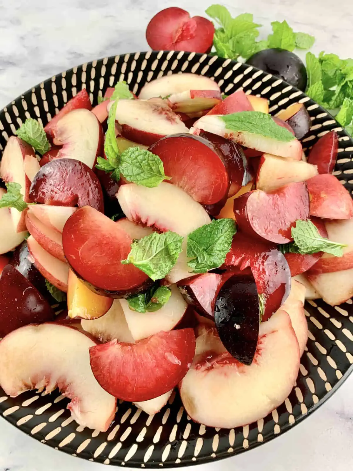 A close-up of white peach salad on a black patterned plate with plums and mint on the side.