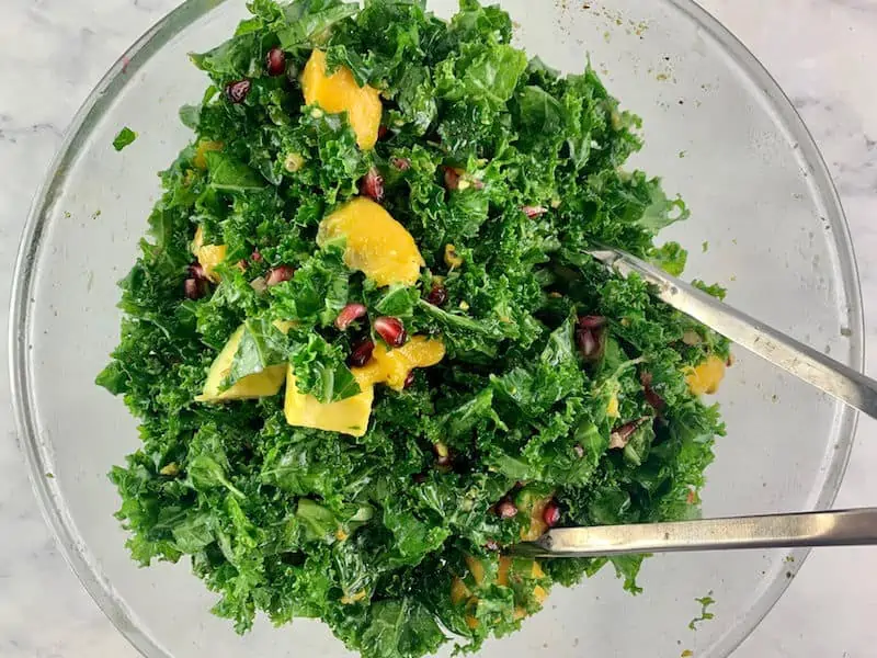 Mixinhpg kale mango salad in a glass bowl with tongs
