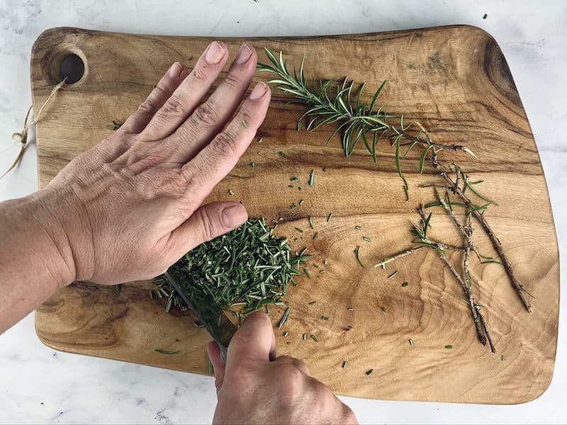 HANDS FINELY CHOPPING ROSEMARY ON WOODEN BOARD WITH KNIFE