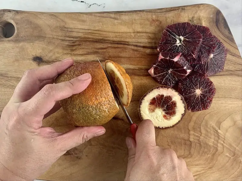 HANDS TRIMMING BLOOD ORANGE ON WOODEN BOARD WITH KNIFE