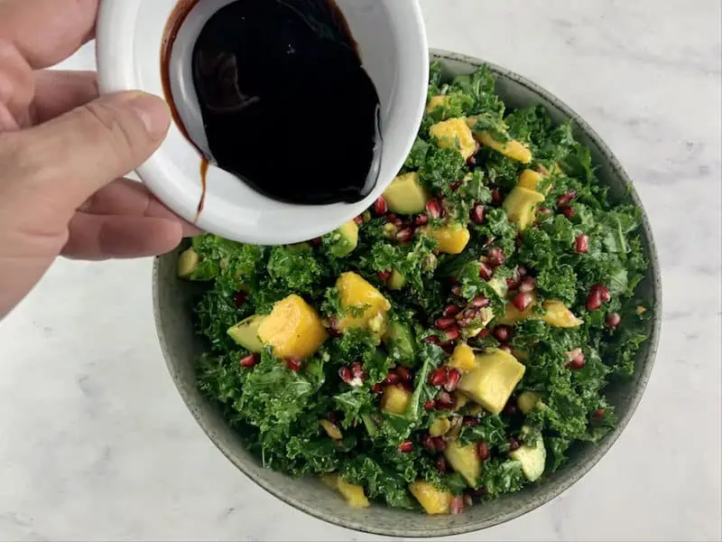 Hands drizzling pomegranate molasses from white container over kale mango salad