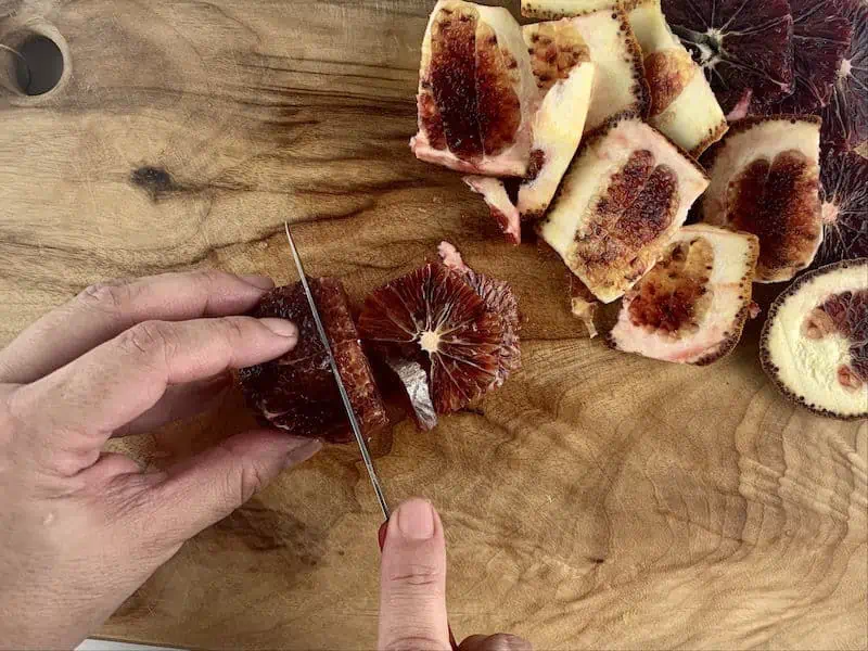 HANDS SLICING BLOOD ORANGES INTO ROUNDS WITH A KNIFE ON WOODEN BOARD, PEEL ON SIDE