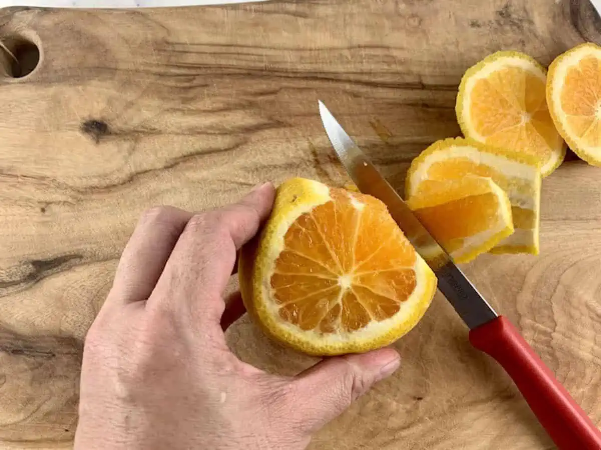 Hands cutting peel from orange on a wooden board with knife.