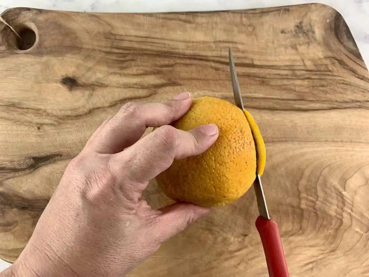 Hands trimming the ends from an orange on a wooden board with a knife.