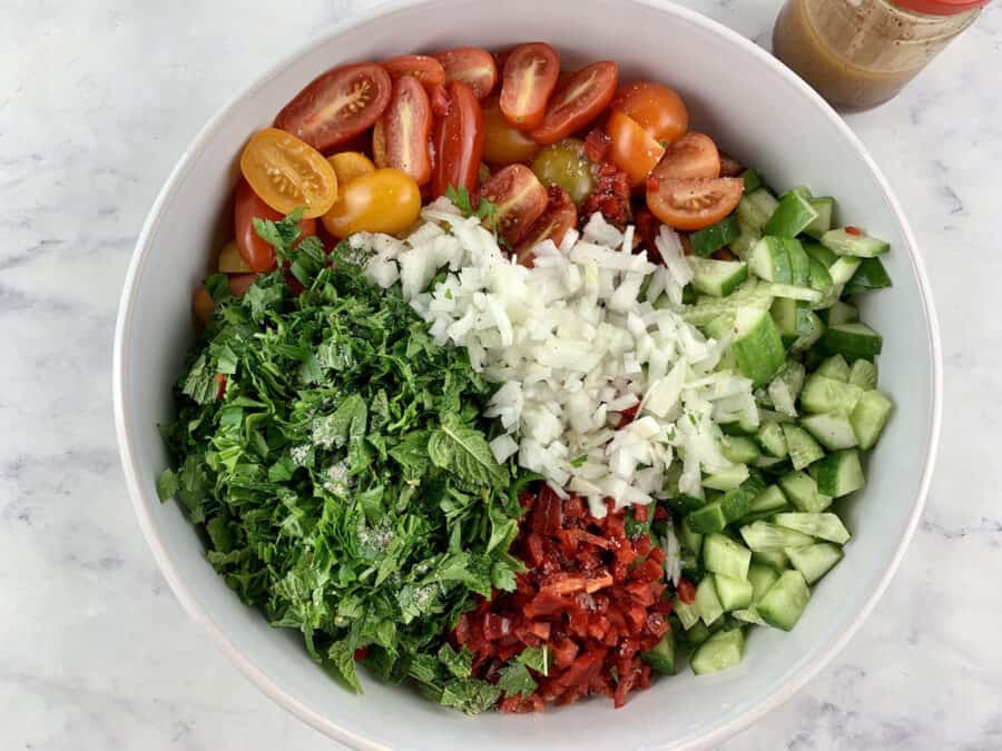 Cut sumac salad ingredients in a white bowl, with dressing in a jar in the right corner.