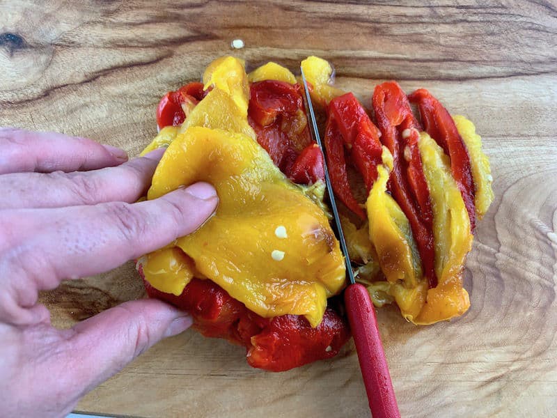 Hands slicing peeled, roasted peppers on a wooden board with a red knife.