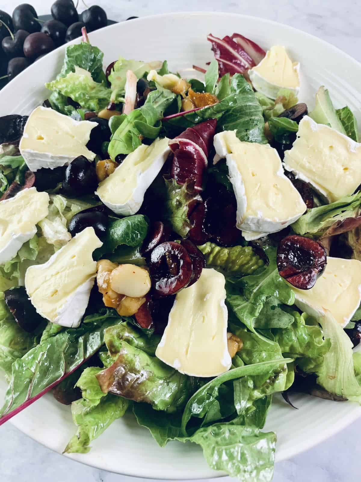 Close up of Brie salad with cherries in a white bowl with cherries in top left corner.